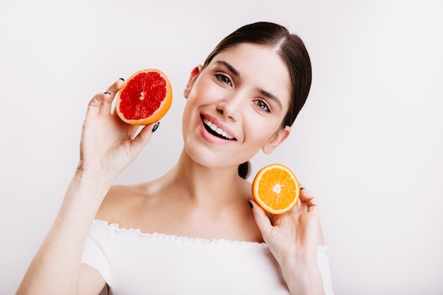 Adorable girl with friendly smile on white wall. Woman without makeup holds slices of juicy orange and grapefruit.