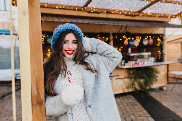 Adorable girl with long brown hair posing with smile near market decorated with christmas garland. Outdoor portrait of joyful european lady in trendy gray coat holding lollipop and laughing.