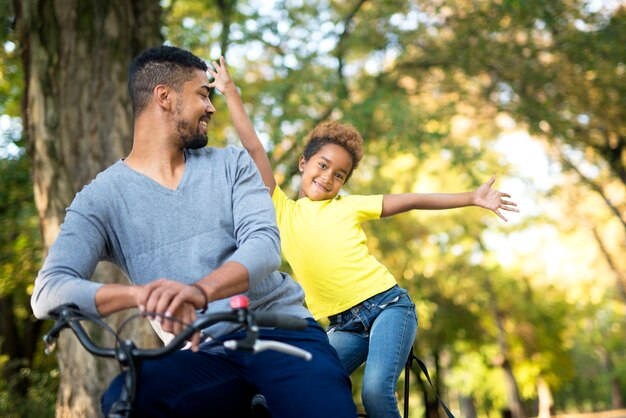 Adorable girl with raised arms and father on a bicycle enjoying in the park