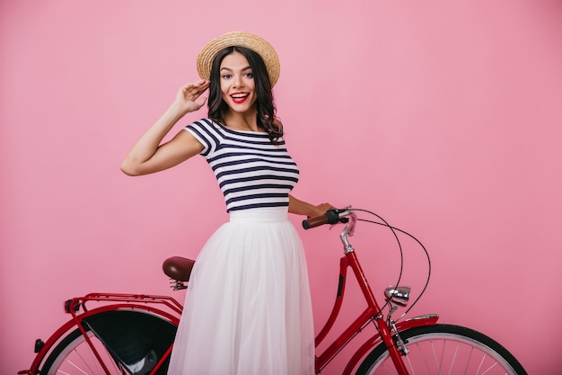 Adorable girl with wavy hair standing near red bicycle. Indoor photo of pleasant slim woman in hat expressing positive emotions.