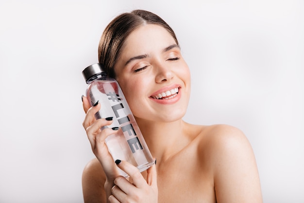 Adorable girl without makeup posing with bottle of water on isolated wall. Smiling model demonstrates importance of water for life.
