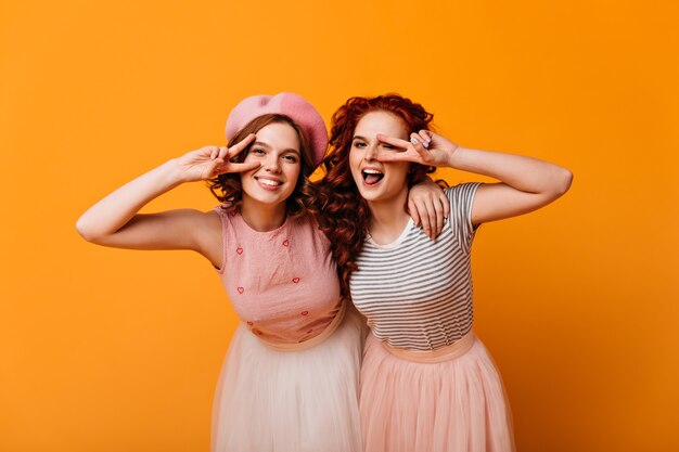 Adorable girls posing with peace signs. Front view of cheerful stylish ladies gesturing on yellow background.