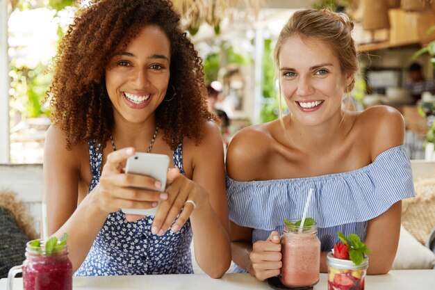 Adorable gorgeous dark skinned female has bushy hairstyle uses smart phone for online communication meets with close friend, recreate at cafeteria with exotic fresh summer cocktails. Leisure concept