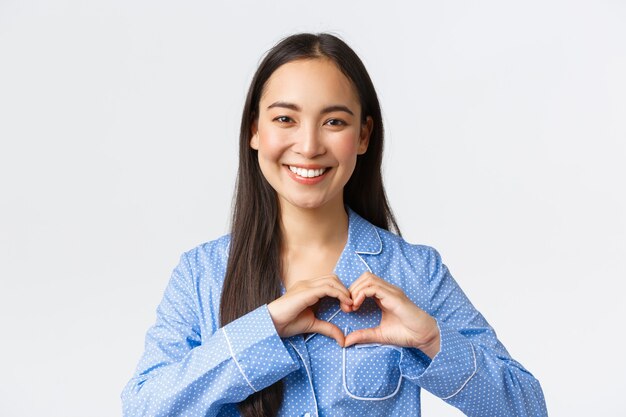 Adorable happy asian girl in blue pajama love staying at home, wearing cozy jammies, showing heart gesture and smiling delighted, standing white background upbeat.