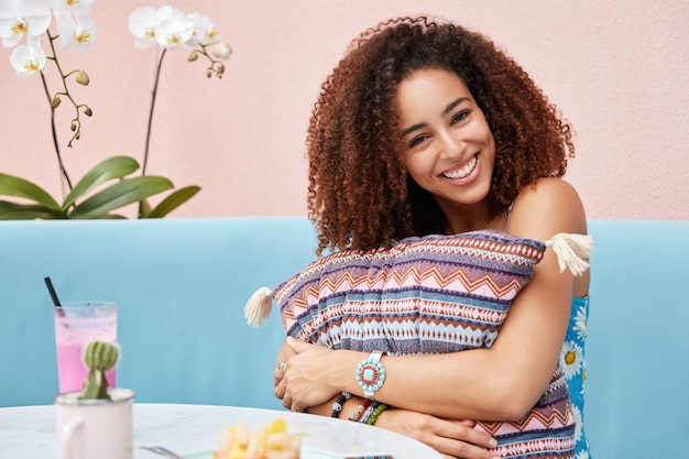 Adorable happy dark skinned female with afro hairstyle, being glad to recieve compliment, embraces cushion, drinks smoothie in cafeteria