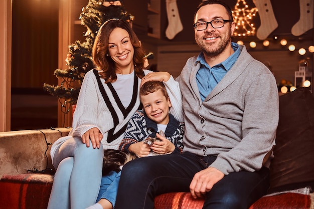 Adorable happy family with their cat hugging together while sitting on sofa in decorated room during Christmas time.