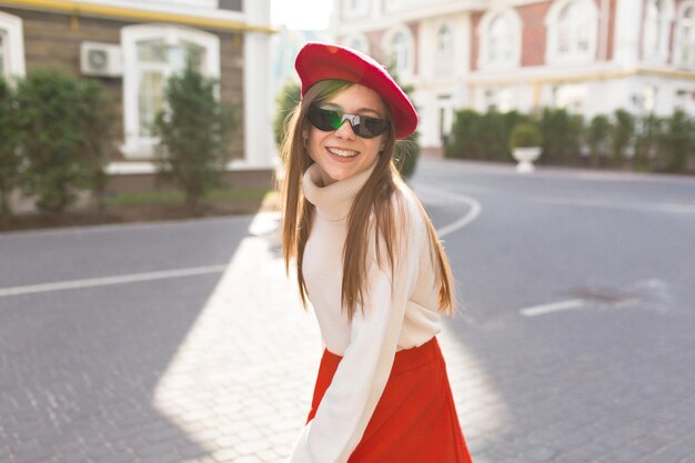 白いプルオーバーとサングラスの赤いベレー帽を身に着けている長い髪の愛らしい幸せな女の子は、カメラを見て、日光に微笑んでいます