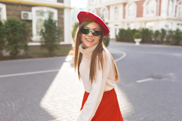 Adorable happy girl with long hair wearing white pullover and red beret in sunglasses is looking at camera and smiling on sunlight