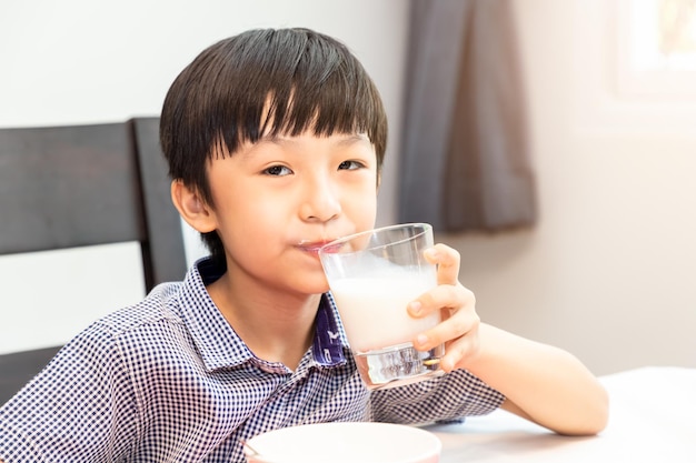 愛らしい幸せな若いアジアの少年が朝食をとり、朝に家でカメラを見て牛乳を飲む