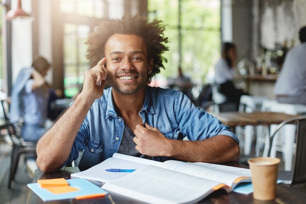 Adorable hipster dark-skinned male student with Afro hairstyle sitting in cafe surrounded with books and copybooks having happy smile while talking by phone