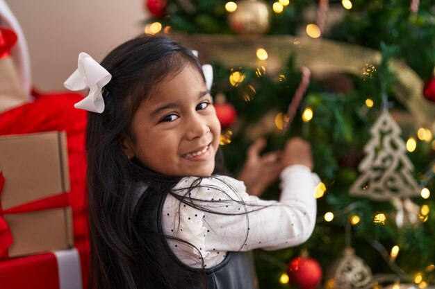 사랑스러운 히스패닉 소녀가 집에서 크리스마스 트리를 장식하며 웃고 있습니다.