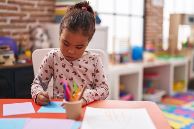 幼稚園で紙に描くテーブルの上に座っている愛らしいヒスパニック系女子学生