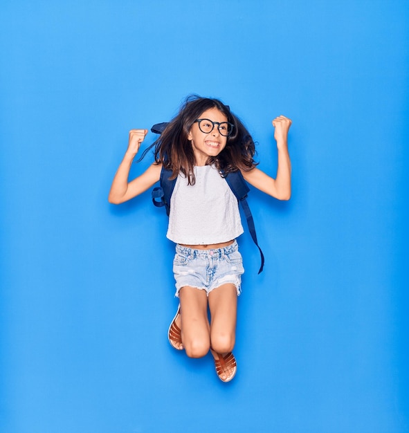 사랑스러운 히스패닉 학생 소녀는 안경을 쓰고 배낭을 메고 행복한 미소를 짓고 고립된 파란색 배경 위에 미소를 지으며 점프합니다