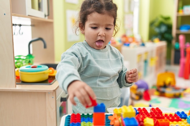 Adorable hispanic toddler playing with construction blocks standing at kindergarten