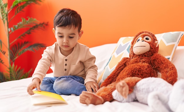 Adorable hispanic toddler reading book sitting on bed at bedroom