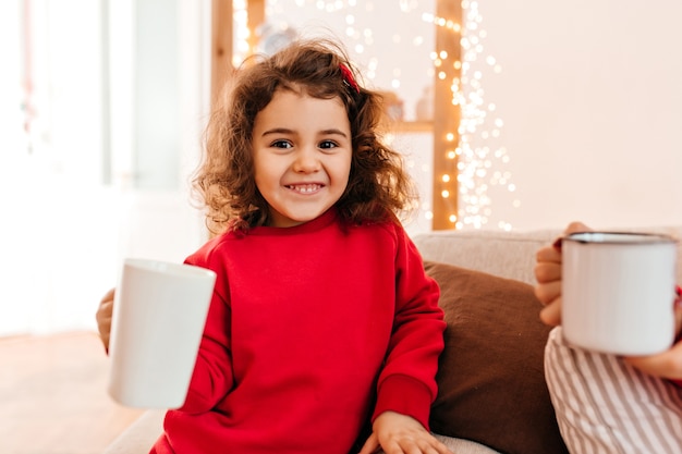 Adorable kid drinking tea. Indoor shot of charming preteen girl in red pajama.