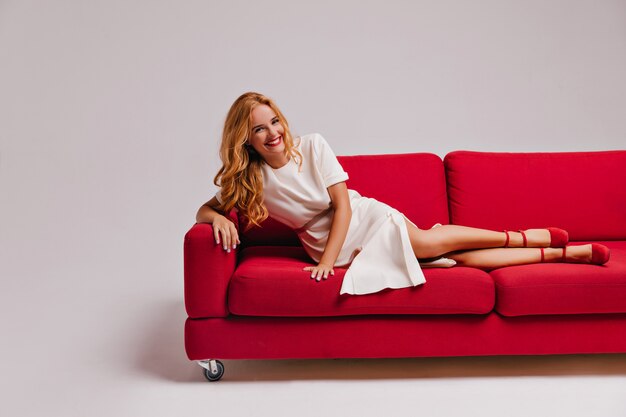 Adorable lady in dress and high heel shoes lying on couch. Refined european girl posing in living room with smile.