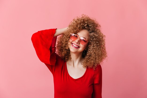 Adorable lady dressed in red blouse laughs while looking at camera. Shot of curly blonde in pink glasses.