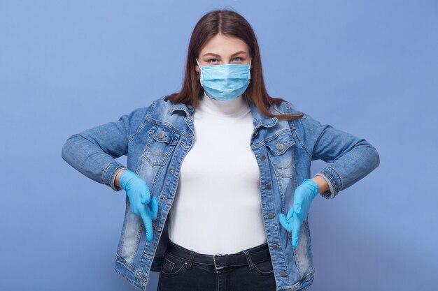 블루 라텍스 장갑과 의료 독감 마스크를 착용하는 동안 검지 손가락을 아래로 가리키는 사랑스러운 아가씨