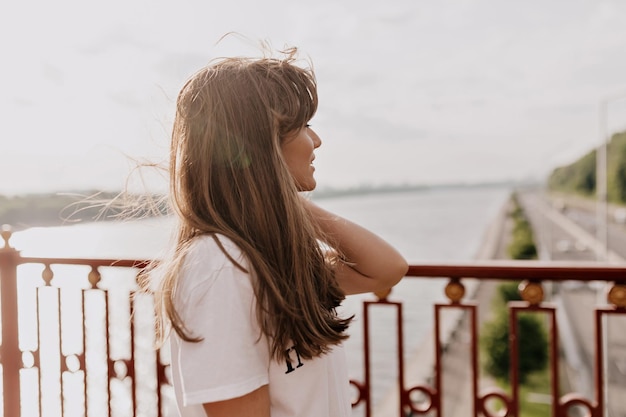 Adorable lady with long dark hair is looking aside on the bridge in morning sunshine Happy european woman walking in the city in warm sunny day