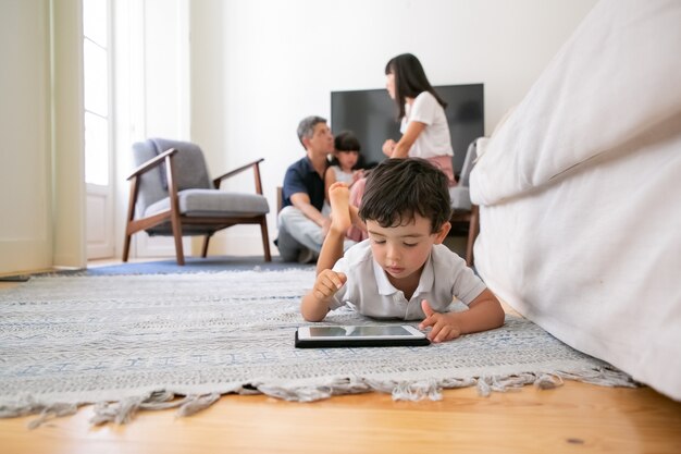 Adorable little boy using tablet, lying on floor in living room while parents and sister sitting together i