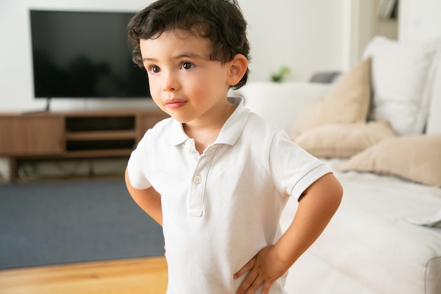 Adorable little boy in white shirt standing with hands on hips in living room