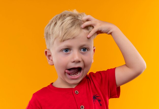 An adorable little boy with blonde hair and blue eyes wearing red t-shirt holding hand on head with open mouth