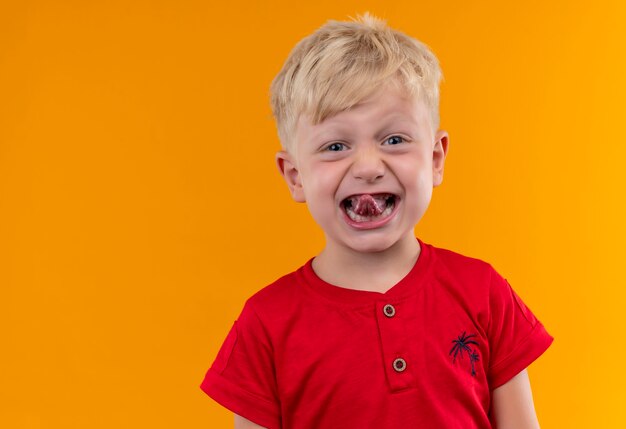 an adorable little boy with blonde hair and blue eyes wearing red t-shirt showing his tongue