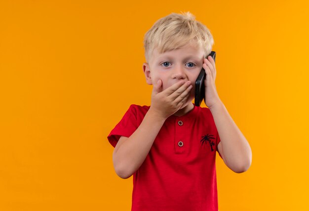 Очаровательный маленький мальчик со светлыми волосами и голубыми глазами в красной футболке разговаривает по мобильному телефону, удивительно глядя, положив руку на рот