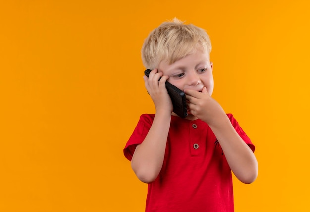 Очаровательный маленький мальчик со светлыми волосами и голубыми глазами в красной футболке разговаривает по мобильному телефону