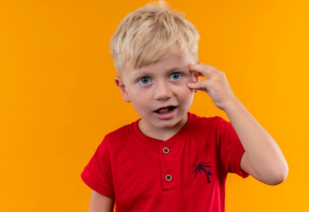금발 머리와 파란 눈을 가진 사랑스러운 어린 소년은 놀라운 빨간 티셔츠를 입고 머리에 손을 대고 있습니다.