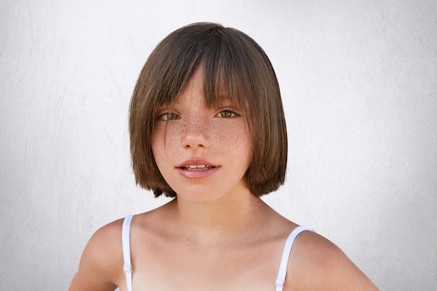 Free photo adorable little child with brown charming eyes, freckled skin and thin lips having stylish hairdo, wearing summer clothes, looking directly into camera while posing on white.