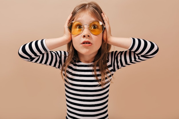 Adorable little cute girl wearing round yellow glasses looks worried, holding hands on the head and looking away