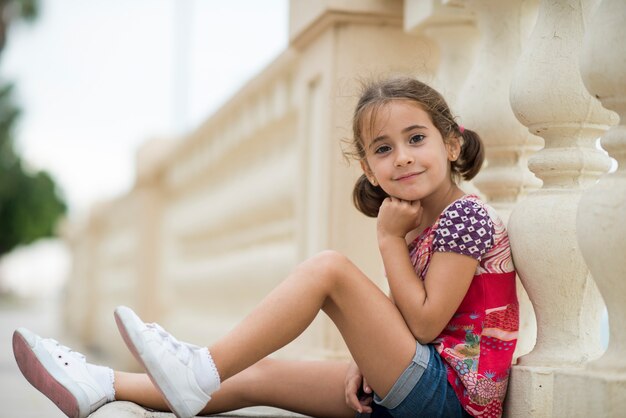 Adorable little girl combed with pigtails outdoors sitting on urban floor.