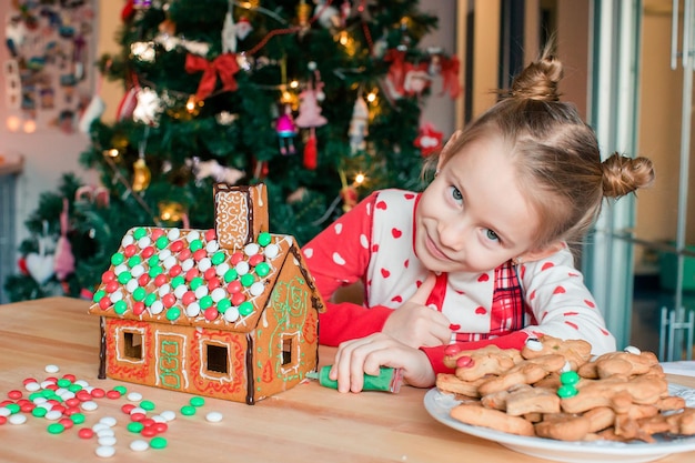 진저브레드 하우스를 조명과 크리스마스 트리의 유약 배경으로 장식하는 사랑스러운 어린 소녀, 양초와 등이 있는 테이블. 휴일을 축하하는 행복한 가족.
