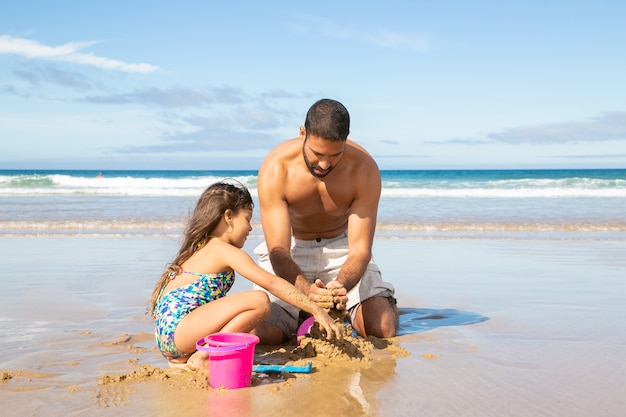 Adorable little girl and her dad building sandcastle on beach, sitting on wet sand, enjoying vacation