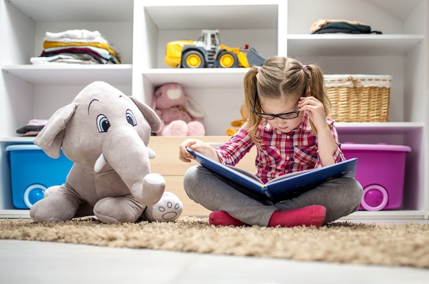 Очаровательная маленькая девочка сидит на ковре и читает книгу для своего плюшевого слона