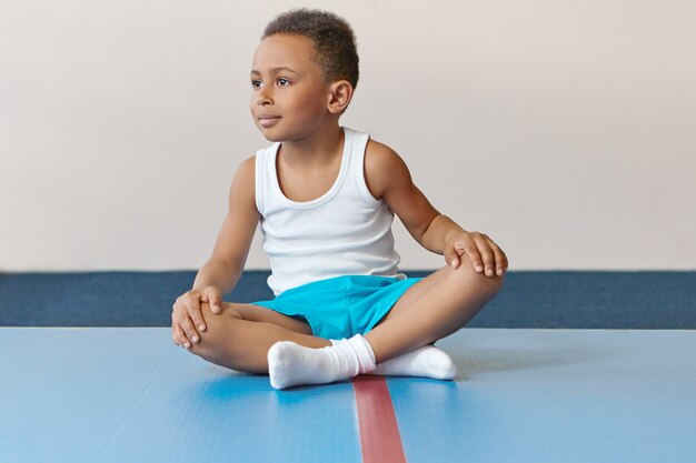 Adorable little sportsman of African appearance sitting on mat with legs crossed relaxing after intensive training.