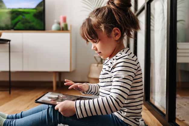 Очаровательная маленькая женщина в хорошем настроении смотрит мультфильмы в планшете и позирует в гостиной.