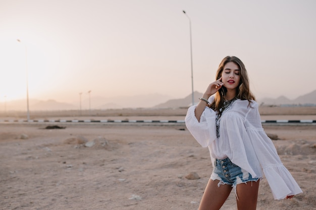 Adorable long-haired woman in vintage blouse posing with eyes closed, touching her face with desert view. Charming pretty young woman in white shirt dancing with pacific face expression