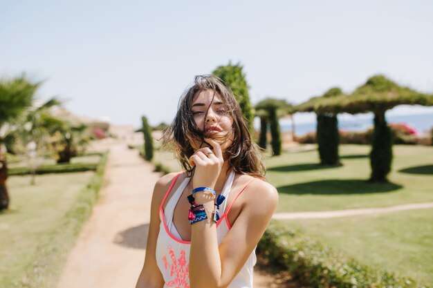 Adorable lovely brunette girl in white tank-top and trendy accessories posing with eyes closed with exotic trees. Portrait of charming tanned young woman spending time outside in morning