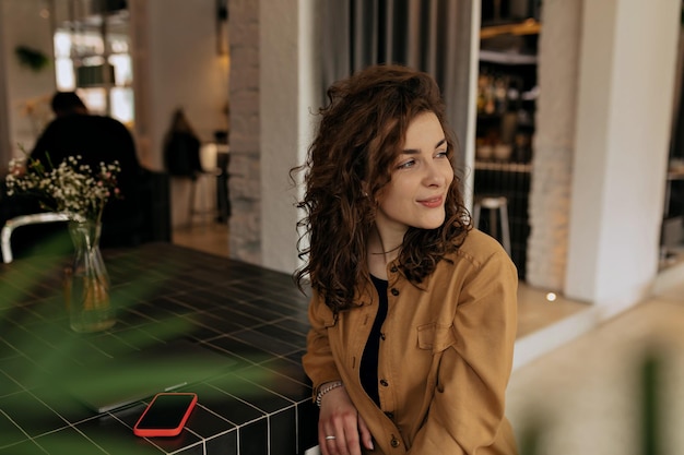 Adorable lovely girls with wavy hair wearing brown shirt is looking aside and smiling Brunette wears casual clothes sitting in cafe Relaxed lifestyle concept