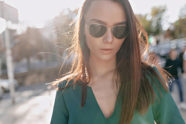 Adorable lovely stylish girl with dark hair in green blouse and sunglasses is posing at camera while walking down the street in sunlight in warm summer day