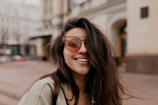 Очаровательная милая женщина с темными волосами в модных очках позирует перед камерой на открытом воздухе во время прогулки по улице
