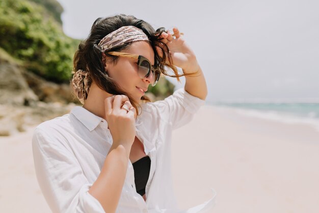 Adorable lovely woman with dark wavy hair dressed white shirt and black sunglasses has fun on the white beach near the ocean with lovely smile.