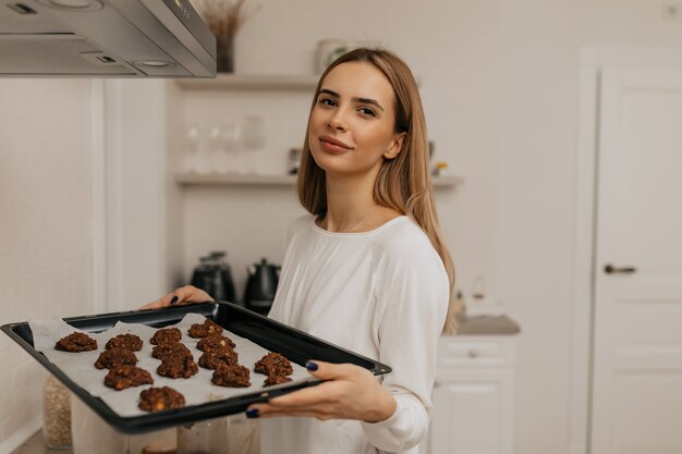 Очаровательная милая женщина со светло-каштановыми волосами в белой рубашке держит деко с печеньем на кухне
