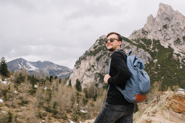 Adorable man wearing sunglasses climbing in mountains and looking away, holding blue backpack