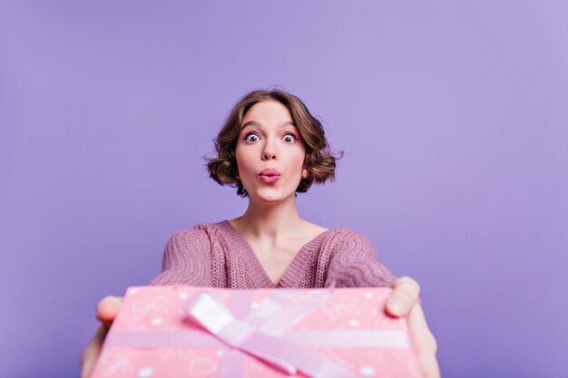 Adorable pale woman posing with kissing face expression and holding birthday gift. interested young lady isolated on purple wall with new year present.