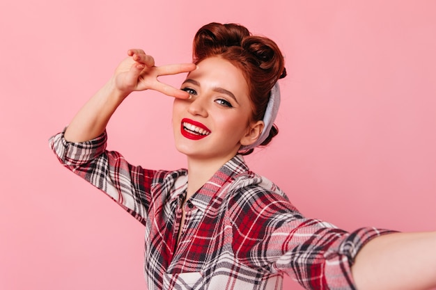 Adorable pinup woman with bright makeup making selfie. Studio shot of appealing girl in checkered shirt showing peace sign.