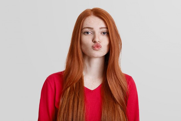 Adorable red haired female with freckled skin, rounds lips, going to kiss someone, has long redish hair, isolated on white. Natural beautiful woman poses indoor. Body language concept